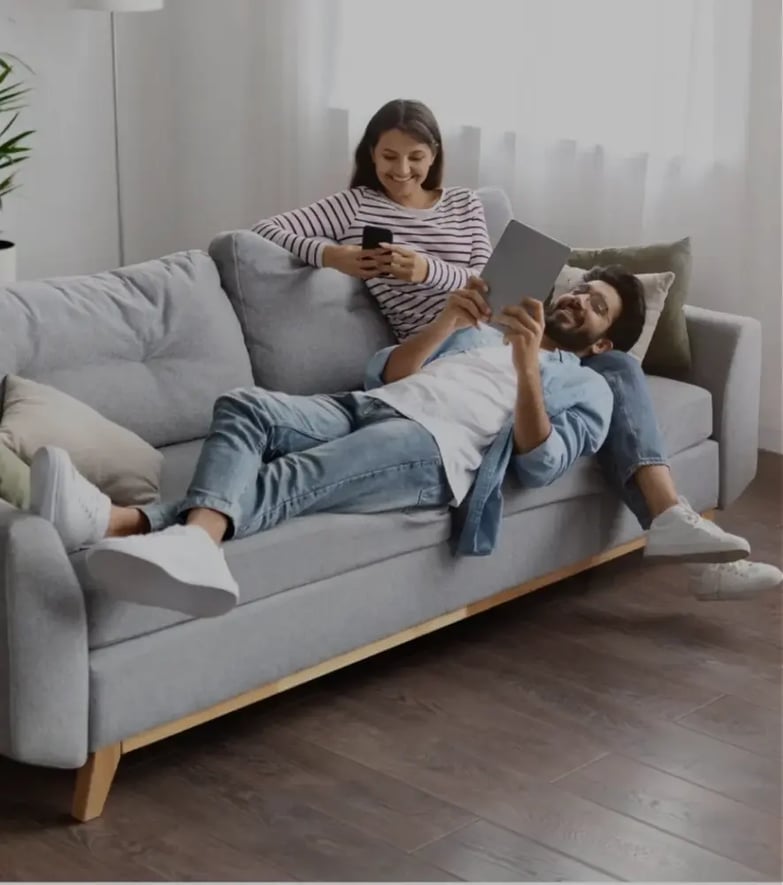 Joven pareja está disfrutando su tiempo juntos, en una sala moderna y amplia. Hay un librero y maceta al fondo, y entra la la luz con amplitud.