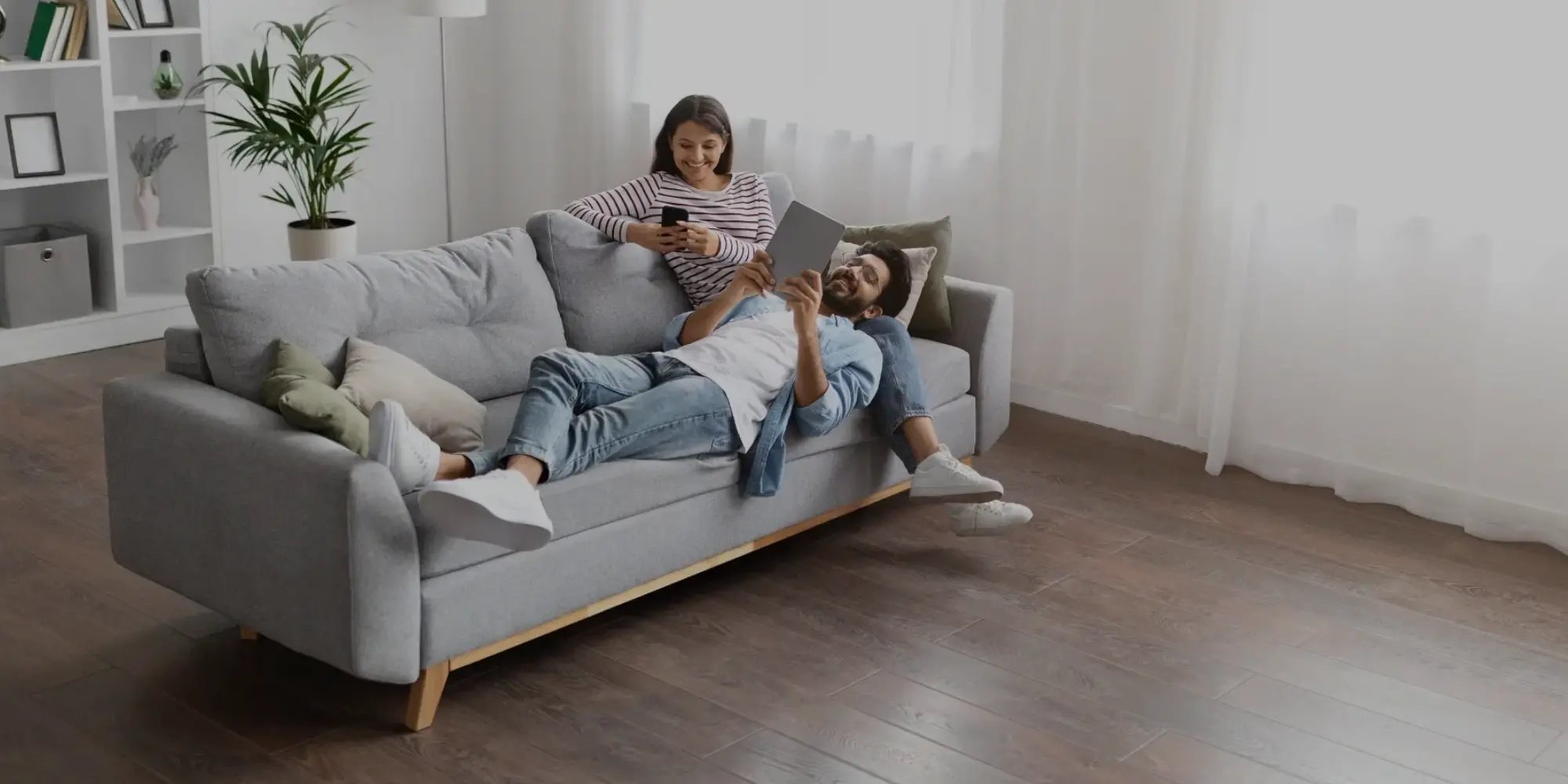 Joven pareja está disfrutando su tiempo juntos, en una sala moderna y amplia. Hay un librero y maceta al fondo, y entra la la luz con amplitud.